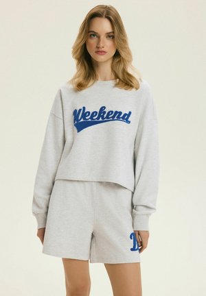 Mladá žena v svetlosivej mikine s nápisom „Weekend“ modrou farbou a v zladených kraťasoch s modrým písmenom „D“ na ľavej nohe.