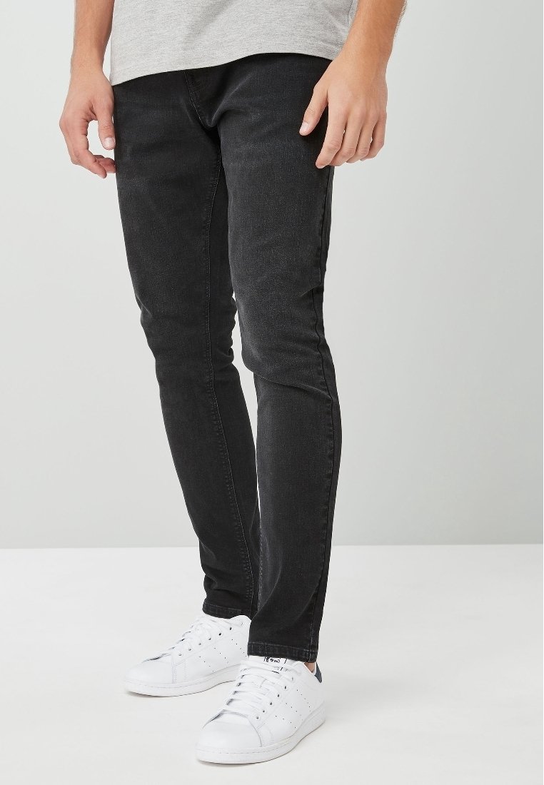 Homme portant un jean noir ajusté, des baskets blanches et un t-shirt gris clair, debout devant un fond gris clair uni.