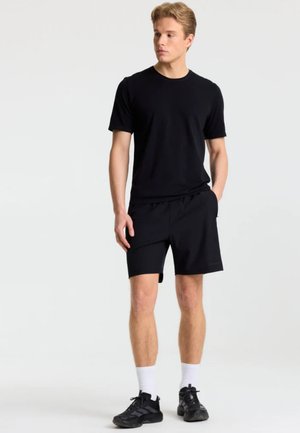 Ung mand iført sort t-shirt, sorte shorts, hvide sokker og sorte sneakers, der står foran en ensfarvet baggrund.