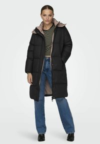 Svart pufferjakke med rosa fôr, til kneet, med glidelås, lommer og hette, kombinert med stripet topp og blå jeans.