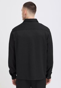 Hombre con cabello corto que lleva una camiseta negra de manga larga, visto desde atrás contra un fondo claro y liso.