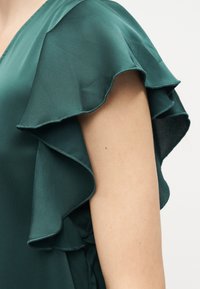 Αμάνικο σκούρο πράσινο μπλούζα με στρωματοποιημένη λεπτομέρεια βολάν στον ώμο, που δείχνει έναν ακάλυπτο άνω βραχίονα σε απλό φόντο.