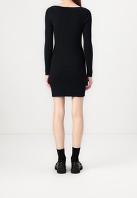Robe en maille côtelée noire avec des manches longues, une silhouette ajustée et un dos dégagé, accompagnée de chaussettes noires et de chaussures noires épaisses.