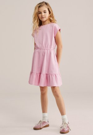 Jong meisje met golvend blond haar, gekleed in een roze jurk met korte mouwen en een gerimpelde zoom, en roze sneakers, staande tegen een neutrale achtergrond.