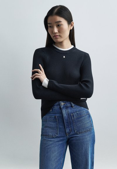 Joven con cabello largo y oscuro, vistiendo un suéter negro acanalado sobre una camisa blanca y jeans azules de tiro alto, con los brazos cruzados, fondo neutro.