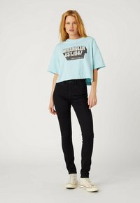 Jasnoniebieski krótki t-shirt z grafiką „WRANGLER EST. 1947”. Połączony z dopasowanymi czarnymi dżinsami i beżowymi trampkami za kostkę. Neutralne tło.