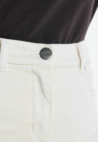 Zbliżenie białych dżinsów z denimu z czarnym guzikiem z napisem "PURE" zapinanym na wysokości talii, noszonych z czarnym topem.