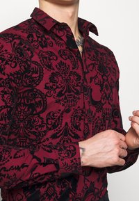 Viininpunainen paita mustalla koristeltulla kukkakuviolla, jossa on kauluskäänne ja pitkät hihat mustilla napituksilla ja kalvosimilla.