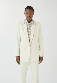 Helle Cremefarbene Anzugjacke mit Revers, zwei aufgesetzten Taschen und einer Knopfschließe. Darunter eine gemusterte Bluse mit Kragen.