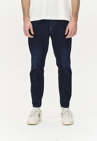 Sötétkék farmernadrág, slim fit szabással, öt zsebbel és diszkrét kopásokkal. Fehér tornacipővel párosítva.