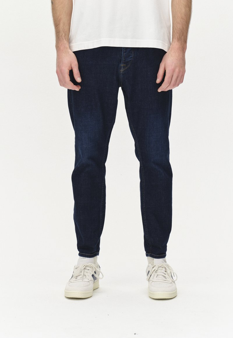 Sötétkék farmernadrág, slim fit szabással, öt zsebbel és diszkrét kopásokkal. Fehér tornacipővel párosítva.