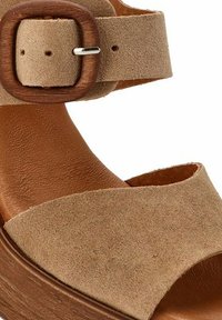 Sandalia de ante beige con una correa ancha y una hebilla ajustable de madera oscura. Presenta una suela de plataforma marrón lisa. Superficie superior texturizada.