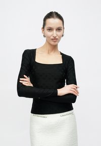 Μαύρο τοπ με μακριά μανίκια, τετράγωνο ντεκολτέ και υφασμάτινη υφή. Η φούστα είναι λευκή με υφή και διακοσμητικές λεπτομέρειες.