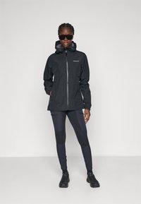 Woman wearing black hooded rain jacket, dark leggings with patches, black sneakers, and sunglasses standing against a plain light background.