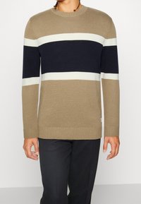 Jasnobrązowy wełniany sweter z granatowymi i kremowymi poziomymi paskami, okrągły dekolt, ściągacze na rękawach i dole. Noszony z ciemnymi spodniami.