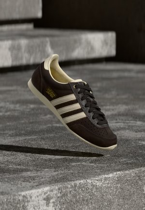 Ciemnobrązowy but Adidas Japan z kremowymi paskami i podeszwą, unoszący się nad betonową powierzchnią z kamiennymi schodami w tle.