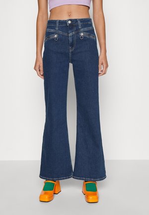 Granatowe, rozszerzane jeansy z wysokim stanem i przednimi kieszeniami na zamek, noszone z jasnopomarańczowymi butami na klocku i zielonymi skarpetkami.