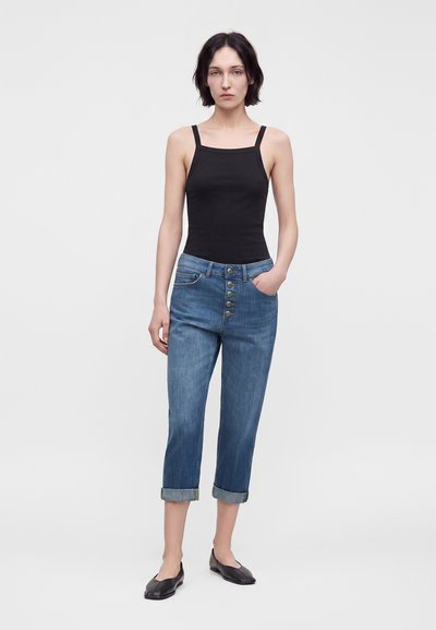 Jeune femme debout portant un haut noir sans manches, un jean bleu retroussé avec boutons apparents, et des chaussures plates noires sur un fond clair uni.