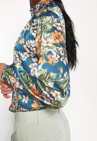 Blouse fleurie en tissu satin, avec un fond bleu, des fleurs multicolores et un col à volants. Manches longues avec poignets froncés.