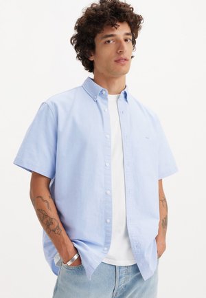lichtblauw overhemd met korte mouwen, met een kraag, witte knopen en een klein logo op de borst, gedragen over een wit T-shirt.