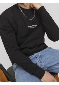 Felpa nera con maniche lunghe, colletto rotondo e stampa bianca "Originals Studio" sul davanti. Indossata con jeans in denim azzurro chiaro.