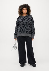 Sweater cinza de malha com padrão de leopardo preto, mangas longas, punhos e bainha canelados. Combinado com calças largas pretas e sapatos pretos robustos.
