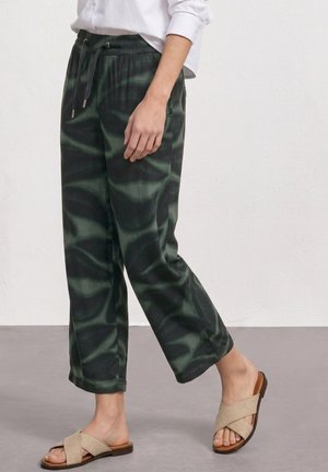 Osoba nosząca ciemnozielone spodnie z nadrukiem tie-dye z sznurkiem i beżowe sandały z krzyżującymi się paskami i odkrytymi palcami, stojąca przy gładkiej jasnej ścianie.