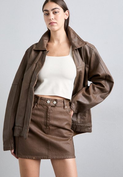 Brun læder oversized jakke med høj krave, parret med en hvid cropped top og en brun denim miniskirt med forlommer og knaplukning.