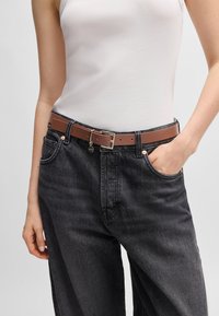 Cintura in pelle marrone con fibbia argentata, indossata sopra jeans in denim grigio scuro. La cintura presenta un piccolo ciondolo appeso vicino alla fibbia.