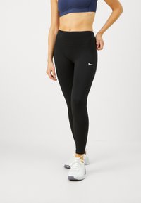 Črne legice z visokim pasom, izdelane iz raztegljivega materiala, z majhnim belim logotipom Nike in belimi stranskimi črtami, v kombinaciji z belimi supergami.