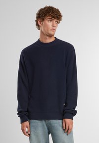 Maglione lavorato a maglia blu navy con texture a coste, scollo rotondo e polsini elasticizzati. Dettaglio del logo visibile all'orlo inferiore.