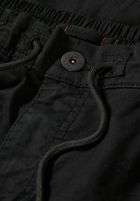 Gros plan sur la ceinture d'un pantalon noir avec un dos élastique, un bouton et un cordon de serrage passé dans des passants cousus.