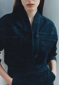 Veste en denim foncé avec col châle, deux poches poitrine et fermeture à boutons sur le devant. Le tissu a une texture subtile et une coupe ajustée.