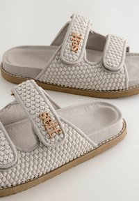 Lys grå slip-on sandaler med struktureret vævet stof, Velcro-stropper og en brun gummisål. Har et logo akcent på stropperne.