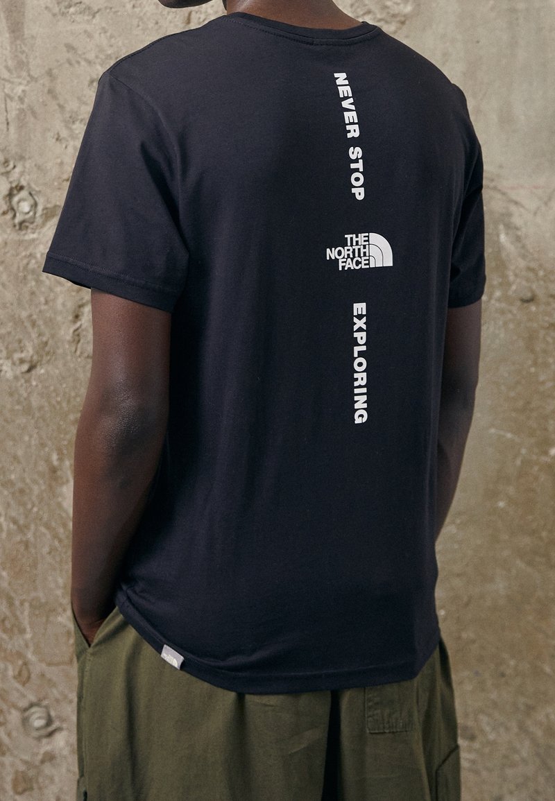 Persona que lleva una camiseta negra de The North Face con "Never Stop Exploring" impreso verticalmente en la parte posterior, combinada con pantalones verdes.