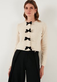 Cardigan di colore crema, morbido e peloso, con maniche lunghe e cinque fiocchi di velluto neri come chiusure sul davanti. Abbinato a pantaloni neri.