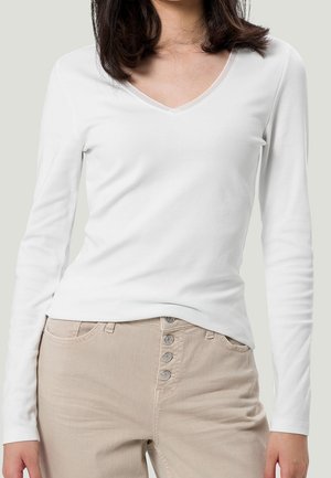 Kobieta ubrana w biały, długi top z dekoltem w serek oraz beżowe spodnie z wysokim stanem, z przodu widoczne cztery guziki.