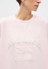 Ανοιχτό ροζ φούτερ με ραμμένο λαιμόκηπο, που διαθέτει κεντημένο κείμενο "ATELIER 2010 MOS MOSH" σε εκρού σε όλο το στήθος.