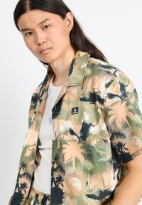 Korte mouwen, gekleed shirt in beige, groen en zwart met een palmboomprint, voorzien van een klein logo-embleem op de linkerborst.