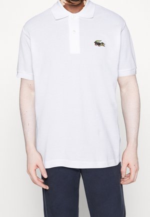 Mann trägt ein weißes kurzärmliges Poloshirt mit kleinem Vogel-Logo auf der linken Brust und dunkle Hose vor einem schlichten Hintergrund.