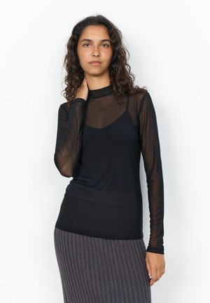 Femme aux cheveux bouclés portant un haut noir transparent à manches longues sur un caraco noir et une jupe grise côtelée, debout sur un fond blanc.