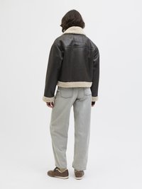 Giacca in finta pelle marrone con colletto in shearling crema, design corto e maniche lunghe, abbinata a pantaloni in denim grigi e sneakers marroni.