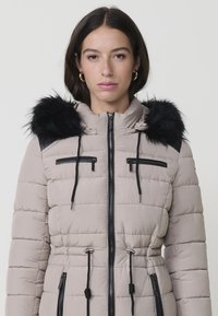 Chaqueta acolchada beige con capucha de piel sintética negra, cintura ajustable, bolsillos con cremallera y detalles negros en los hombros y cremalleras.