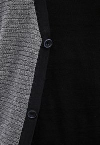 Svart och grå stickad cardigan med ribbat mönster på den grå sidan. Har en rund svart knapp för stängning på framsidan.