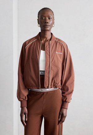 Femme portant une veste marron à fermeture éclair et un pantalon assorti avec un haut de sport blanc, posant devant un fond gris clair uni.