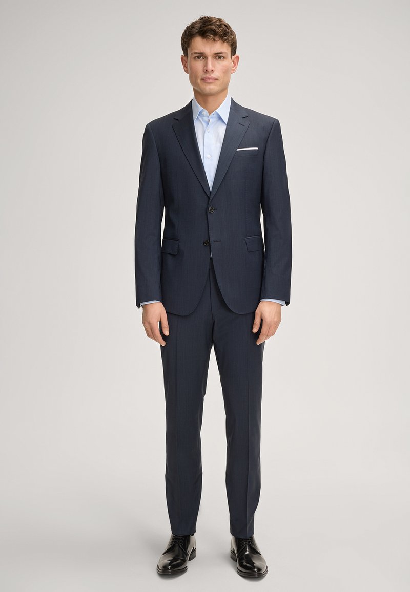 Granatowy garnitur o fakturowanym wykończeniu, jednorzędowy, z dwoma guzikami i klapami wyciętymi, w połączeniu z jasnoniebieską koszulą i czarnymi butami.
