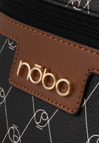 Zwarte gestructureerde stoffen tas met een wit geometrisch patroon, bruin leren accent en een goudkleurig logo op een rechthoekig label.