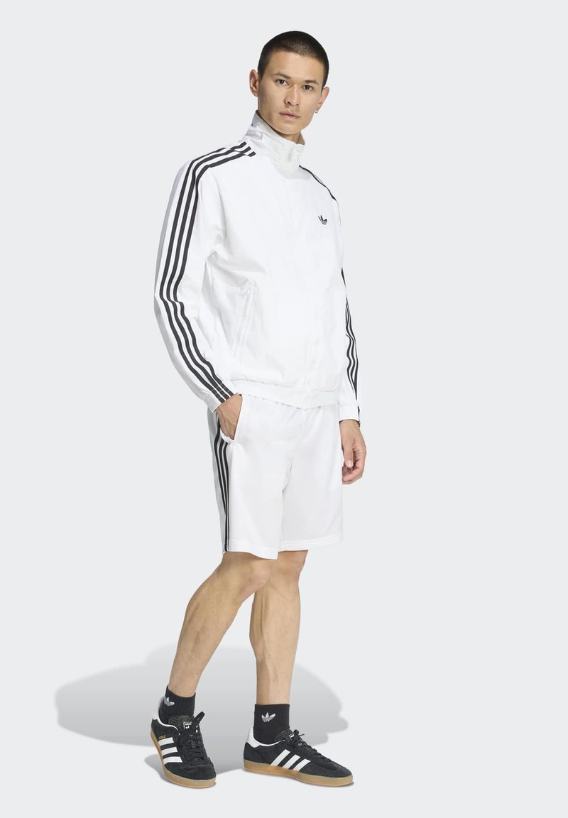 Uomo che indossa giacca e pantaloncini della tuta Adidas bianchi con strisce nere, calzini Adidas neri e scarpe da ginnastica nere con strisce bianche.