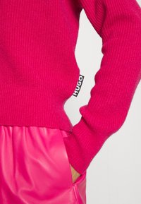 Magenta prążkowany sweter dziany z dopasowaną talią, zestawiony z błyszczącymi różowymi spodniami ze sztucznej skóry. Po boku znajduje się czarna metka z napisem "HUGO".