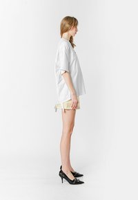 Camisa blanca de gran tamaño con mangas cortas, combinada con pantalones cortos amarillos claros. Tacones negros con un acento decorativo. Vista de perfil lateral.